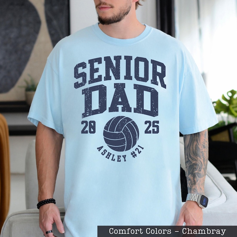 A man wearing a light blue t-shirt with a graphic design that says "SENIOR DAD" and "ASHLEY #21" printed on it.