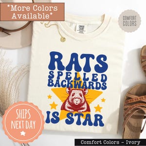 May include: Ivory Comfort Colors t-shirt with the text "RATS SPELLED BACKWARDS IS STAR" in blue and white. A graphic of a rat is centered on a yellow star. The shirt has a crew neck and short sleeves.