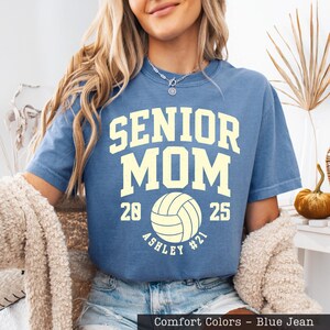 A woman wearing a blue t-shirt with the text "SENIOR MOM" printed on it, along with a volleyball graphic. She is also wearing a necklace and has long blonde hair.