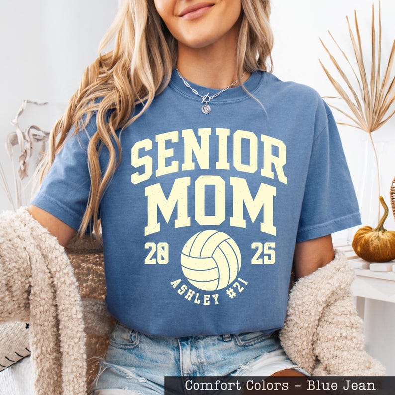 A woman wearing a blue t-shirt with the text "SENIOR MOM" printed on it, along with a volleyball graphic. She is also wearing a necklace and has long blonde hair.