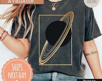 May include: A charcoal gray short-sleeve t-shirt featuring a graphic print of a black planet with gold rings enclosed within a thin gold rectangular frame. The shirt has a crew neckline and a washed, vintage-inspired texture throughout the fabric.