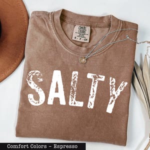 A brown t-shirt with the word "SALTY" printed in white letters, along with a necklace and a hat, all laid out on a white surface.