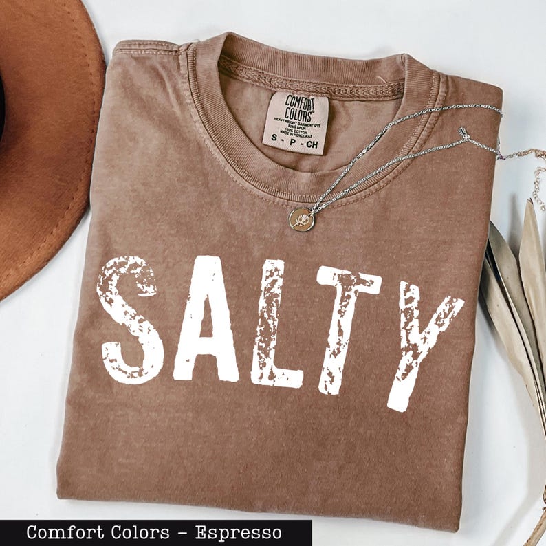 A brown t-shirt with the word "SALTY" printed in white letters, along with a necklace and a hat, all laid out on a white surface.