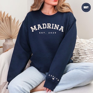 A woman wearing a navy blue sweatshirt with the text "MADRINA EST. 2025" printed on it, sitting on a couch.