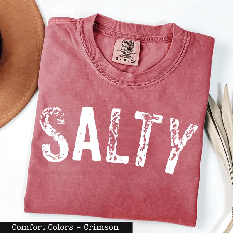 A red t-shirt with the word "SALTY" printed on it, along with a brown hat and some leaves, all laid out on a white surface.