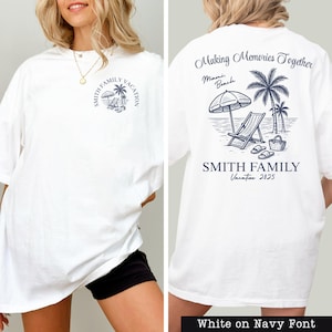A white t-shirt with a graphic design on the back, featuring a beach scene with a palm tree, beach chair, and umbrella.