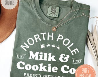 May include: An olive green short-sleeve t-shirt featuring white text that reads North Pole Milk & Cookie Co. Est. 1802 Baking Fresh Daily, with a graphic of Santa in a sleigh pulled by reindeer. The shirt has a Comfort Colors brand neck label.