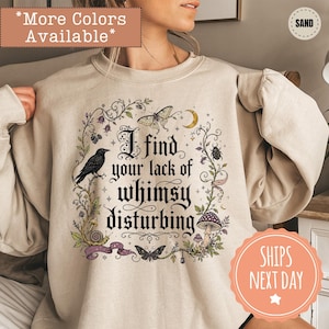 May include: A sand-colored crewneck sweatshirt featuring a graphic print of a raven, moths, a snail, a mushroom, and a crescent moon surrounded by vines. The text reads, I find your lack of whimsy disturbing, in a black gothic-style font.