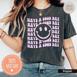May include: A black t-shirt with a pink retro-style smiley face and the text "Have a Good Day" repeated in a wavy font.