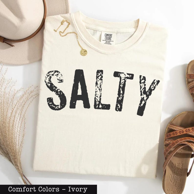 A white t-shirt with the word "SALTY" printed on it, along with a beige hat, a gold necklace, and a pair of sandals, all laid out on a white background.