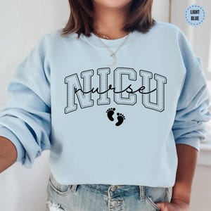 A woman wearing a light blue sweatshirt with the word "NICU" printed on it, along with a pair of jeans.