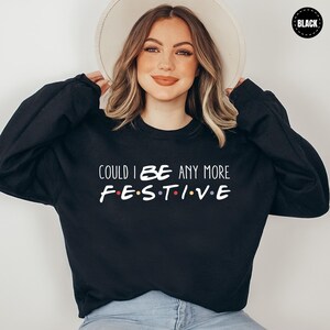 A woman wearing a black sweater with the text "COULD I BE ANY MORE FESTIVE" printed on it, smiling and posing with her hands behind her head.