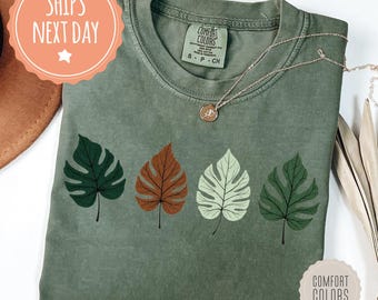 May include: A moss green Comfort Colors t-shirt featuring four stylized leaf designs in shades of green, brown, and white. A delicate gold necklace rests on the shirt. The shirt has a tag that says "Comfort Colors".