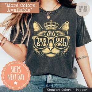 May include: A charcoal gray t-shirt featuring a graphic of a cat wearing a crown and sunglasses. The text inside the sunglasses reads THIS IS AN OUT RAGE! The shirt has a short-sleeved crew neck design and a vintage-style washed fabric appearance.