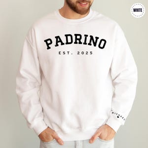 A man wearing a white sweatshirt with the text "PADRINO EST. 2025" printed on it.