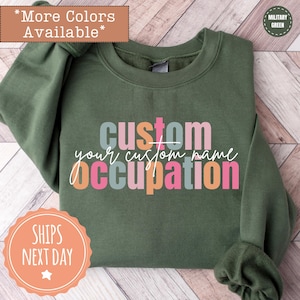 May include: A military green sweatshirt with the words "custom", "your custom name", and "occupation" in a colorful, layered design. The sweatshirt has a crew neck and long sleeves. Additional text includes "More Colors Available" and "Ships Next Day".