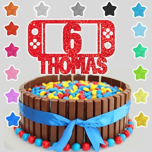 以下が含まれることがあります： ビデオゲームコントローラーのデザイン、数字の6、そして名前のトーマスが書かれた赤いグリッターのケーキトッパー。ケーキトッパーは、カラフルなキャンディーが載ったチョコレートケーキの上に置かれています。
