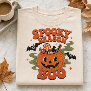 Op de afbeelding: Een crèmekleurig t-shirt met een afbeelding van een oranje pompoen gevuld met snoep. De tekst "SPOOKY SEASON" staat boven de pompoen en "BOO" eronder. Twee zwarte vleermuizen en groene rookaccenten omringen het centrale ontwerp.