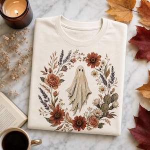 Op de afbeelding: Een crèmekleurig t-shirt met een centrale afbeelding van een witte geest omringd door een cirkelvormige krans van herfstkleurige bloemen, waaronder roestkleurige madeliefjes, rozen, lavendeltakjes en kleine groene cactussen in botanische stijl.