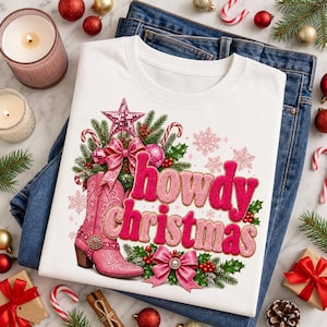 Op de afbeelding: Een wit t-shirt met een afbeelding van een roze cowboylaars, een roze ster en roze strikken omringd door groene dennentakken en rode bessen. De tekst howdy christmas is gedrukt in een roze lettertype met een lichtroze omlijning.