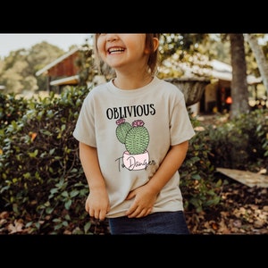 May include: A light beige t-shirt with a cactus graphic and the text "Oblivious To Danger".