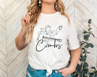 May include: White t-shirt with a line drawing of a country music singer holding a guitar and a cup. The singer is wearing a hat and has a microphone in front of them. The text "Combs" is written in cursive below the drawing.