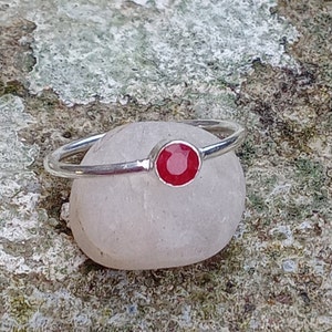 Op de afbeelding: Een zilveren ring met een ronde, helderrode edelsteen. De ring heeft een slanke, gepolijste band en de edelsteen is gezet in een eenvoudige bezel. De ring wordt getoond op een gladde, lichtgekleurde steen tegen een getextureerde achtergrond.
