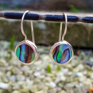Könnte beinhalten: Ein Paar silberne Ohrhänger mit runden, irisierenden Abalone-Muscheleinsätzen. Die Muscheln zeigen eine Mischung aus grünen, blauen und braunen Farbtönen. Die Ohrringe sind an einfachen, gebogenen Silberhaken aufgehängt. Der Hintergrund ist unscharf.