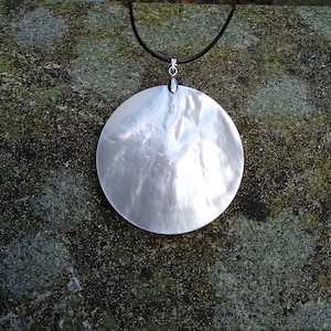 Puede incluir: Un collar con un colgante redondo de concha blanca en un cordón negro. El colgante es grande y tiene una superficie lisa e iridiscente.