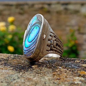 Könnte beinhalten: Ein Silberring mit einer länglichen, ovalen Einlage aus irisierendem Abalone-Muschelmaterial in leuchtenden Blau- und Grüntönen. Die Metallschiene weist an den Seiten ein texturiertes, geätztes Muster auf, das in eine glatte, polierte Silberfassung übergeht.