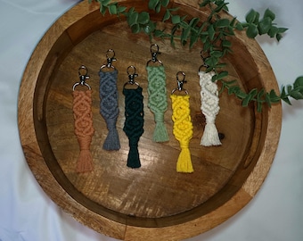 May include: Six colourful macrame keychains with silver key rings. The keychains are displayed on a wooden tray with green foliage.