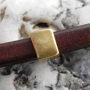 May include: A close-up of a brown leather strap with a gold metal clasp. The clasp is rectangular and has a smooth surface.