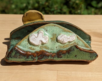 May include: A ceramic dish shaped like a green hill with a yellow sun and white clouds. The dish has a rustic, handmade look.