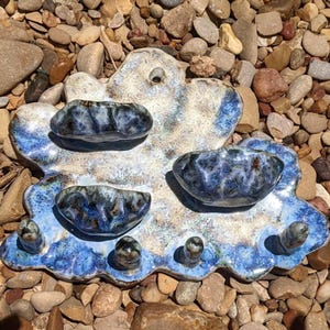 May include: A ceramic wall-mounted organizer featuring a cloud-like shape with a mottled white and blue glaze. It includes three small attached bowls for storage and four protruding pegs at the bottom for hanging items, with a hole for mounting.
