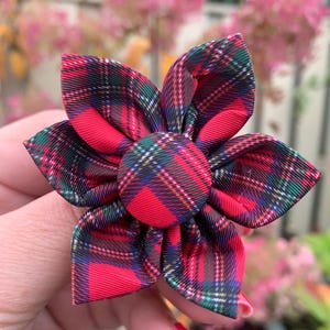 May include: A tartan fabric flower with six petals, predominantly red and green. The central disc matches the plaid pattern. The flower is held in a hand, with a blurred backdrop of pink flowers and foliage.