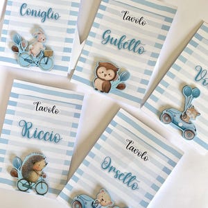 Può includere: Diversi segnaposto con sfondi a righe blu e bianche. Ogni biglietto presenta un'illustrazione di animale diversa e un nome in italiano. I biglietti raffigurano un coniglietto in bicicletta, un gufo, un riccio, un orso in macchina e una volpe in macchina.