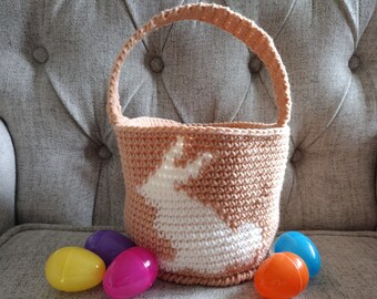 May include: A crocheted Easter basket with a white bunny silhouette design. The basket is peach colored and is sitting on a gray couch with four colorful plastic Easter eggs in front of it.