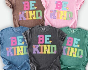 May include: Five Comfort Colors t-shirts in various colors, each featuring the words "BE KIND" in colorful block letters with star accents. The shirts are in shades of brown, pink, blue, charcoal gray, and green.