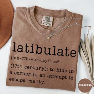 May include: Espresso-colored t-shirt with the word "latibulate" printed in black, along with its definition. A silver necklace with a pendant rests on the shirt. The shirt is a Comfort Colors brand.
