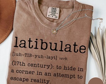 May include: Espresso-colored t-shirt with the word "latibulate" printed in black, along with its definition. A silver necklace with a pendant rests on the shirt. The shirt is a Comfort Colors brand.