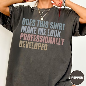 Puede incluir: Una camiseta de manga corta gris carb&oacute;n con el texto desgastado "DOES THIS SHIRT MAKE ME LOOK PROFESSIONALLY DEVELOPED" en l&iacute;neas apiladas de color azul claro, rosa y beige. La prenda tiene cuello redondo y un corte holgado.