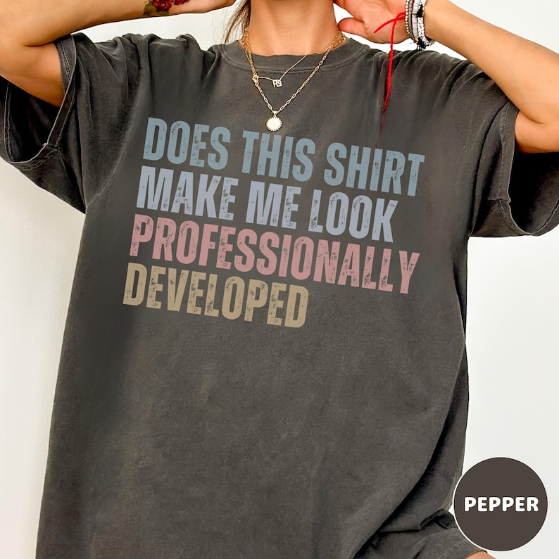 Puede incluir: Una camiseta de manga corta gris carb&oacute;n con el texto desgastado "DOES THIS SHIRT MAKE ME LOOK PROFESSIONALLY DEVELOPED" en l&iacute;neas apiladas de color azul claro, rosa y beige. La prenda tiene cuello redondo y un corte holgado.
