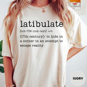 May include: Ivory-colored t-shirt with the word "latibulate" printed in black, along with its pronunciation and definition. The definition reads: "to hide in a corner in an attempt to escape reality."