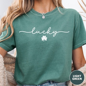 May include: Light green t-shirt with the word "lucky" in white script and a white shamrock. The shirt has short sleeves and a crew neck. The shirt is a solid color.