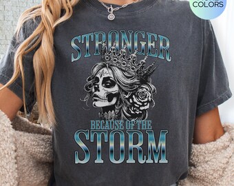 May include: A gray t-shirt with a graphic of a woman with a crown and skull face. The text "Stronger Because of the Storm" is printed on the shirt.