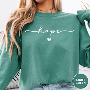 May include: A light green sweatshirt with the word "hope" in a white script font, and a small white heart below the word. The sweatshirt is a solid color and has long sleeves. The words "LIGHT GREEN" are in a white font in a green circle.