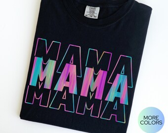 May include: A black short-sleeved t-shirt featuring the word MAMA printed three times in a stacked, overlapping design. The text uses a vibrant, multi-colored gradient effect with shades of pink, purple, and blue. The shirt has a Comfort Colors label.