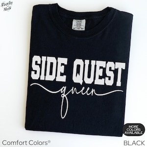 May include: Black t-shirt with the words "SIDE QUEST queen" in white block letters and script. The shirt is a Comfort Colors brand, with a tag at the neckline. The words "MORE COLORS AVAILABLE" and the word "BLACK" are at the bottom right.