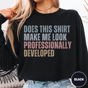 Puede incluir: Una sudadera negra de manga larga con el texto "DOES THIS SHIRT MAKE ME LOOK PROFESSIONALLY DEVELOPED" impreso en una fuente desgastada. Las palabras est&aacute;n coloreadas en azul claro, rosa claro y amarillo claro sobre la tela negra.