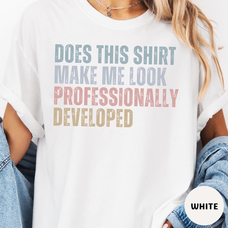 Puede incluir: Una camiseta blanca de manga corta con el texto desgastado "DOES THIS SHIRT MAKE ME LOOK PROFESSIONALLY DEVELOPED" en l&iacute;neas apiladas de colores azul, rosa y beige apagados. La camiseta tiene cuello redondo y un corte holgado.